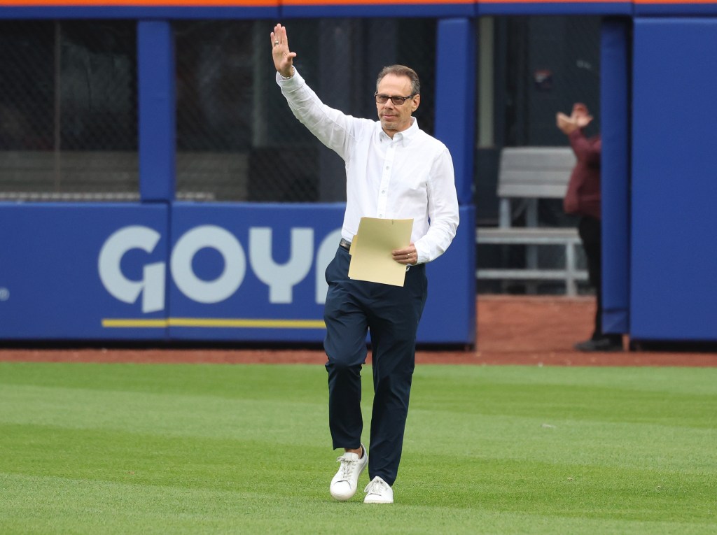 A man in a white shirt and dark pants waves as he walks across a baseball field.