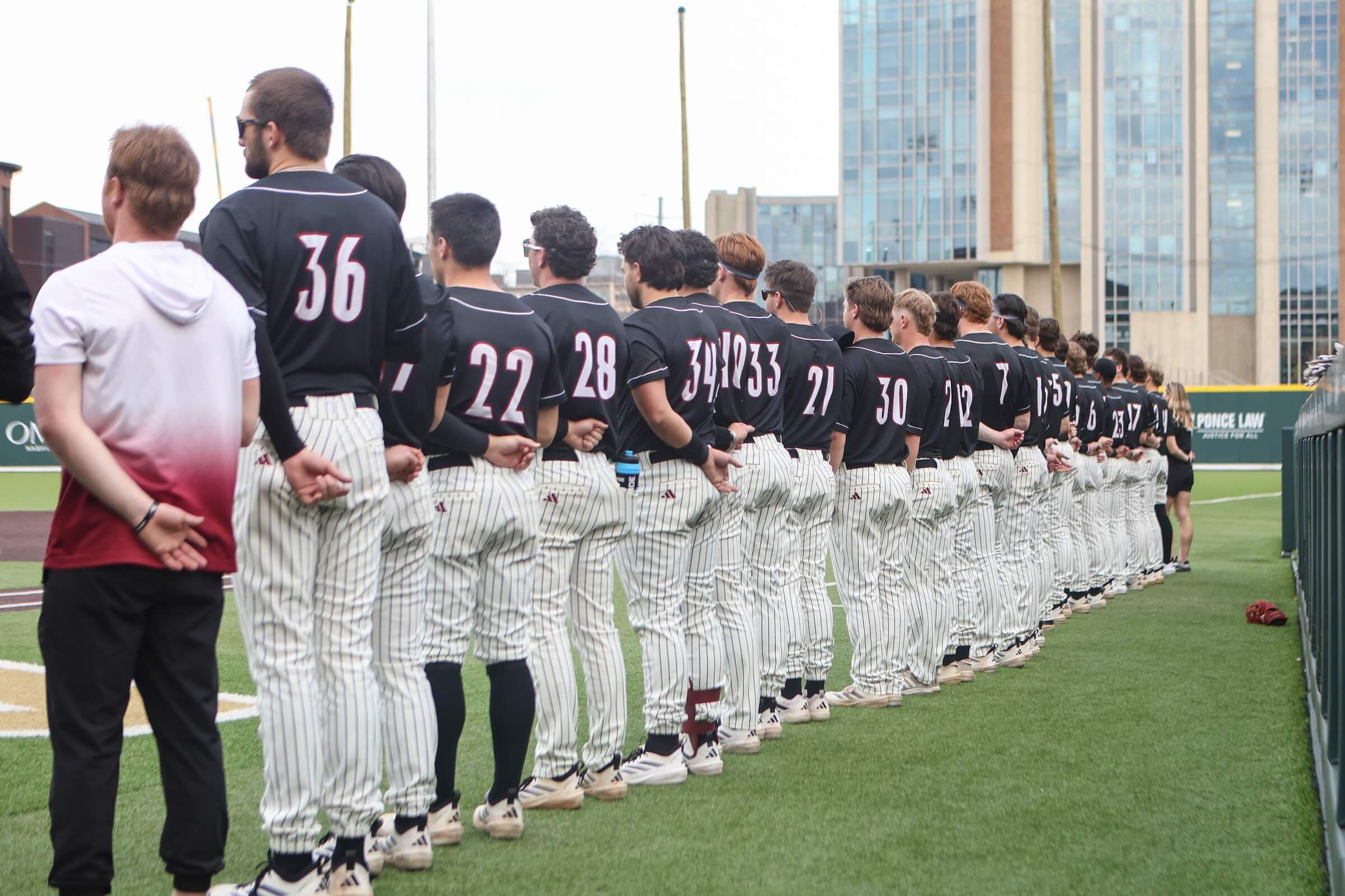 Troy players, wearing their navy jerseys and white pinstripe pants, stand along the first-base line for the national anthem.