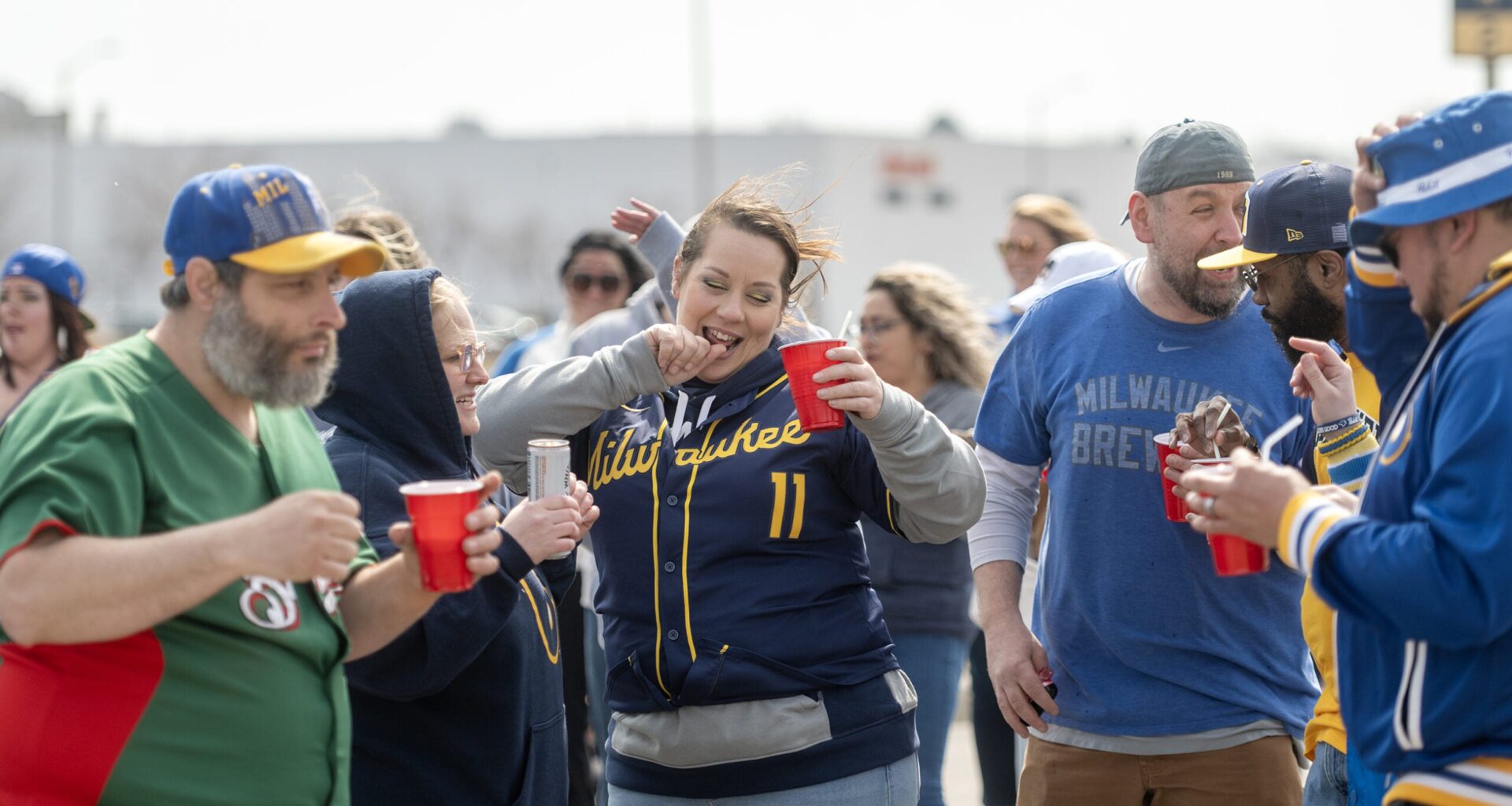 A group of people wearing Milwaukee Brewers apparel gather outdoors, holding red cups and socializing before a game.