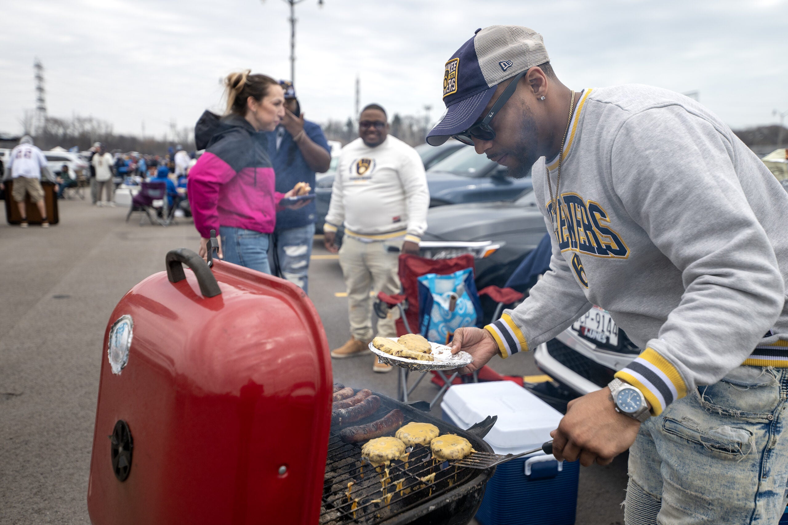 A man grills burgers and sausages on a red barbecue in a parking lot while others stand nearby, tailgating before an event.