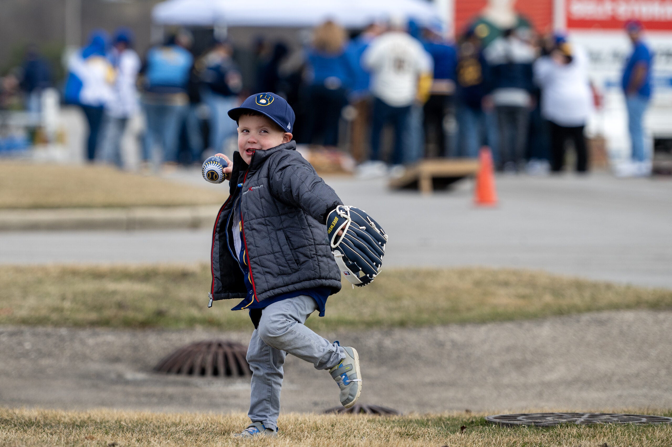 A young boy wearing a baseball glove and cap throws a ball on a grassy area, with a group of people gathered in the background.