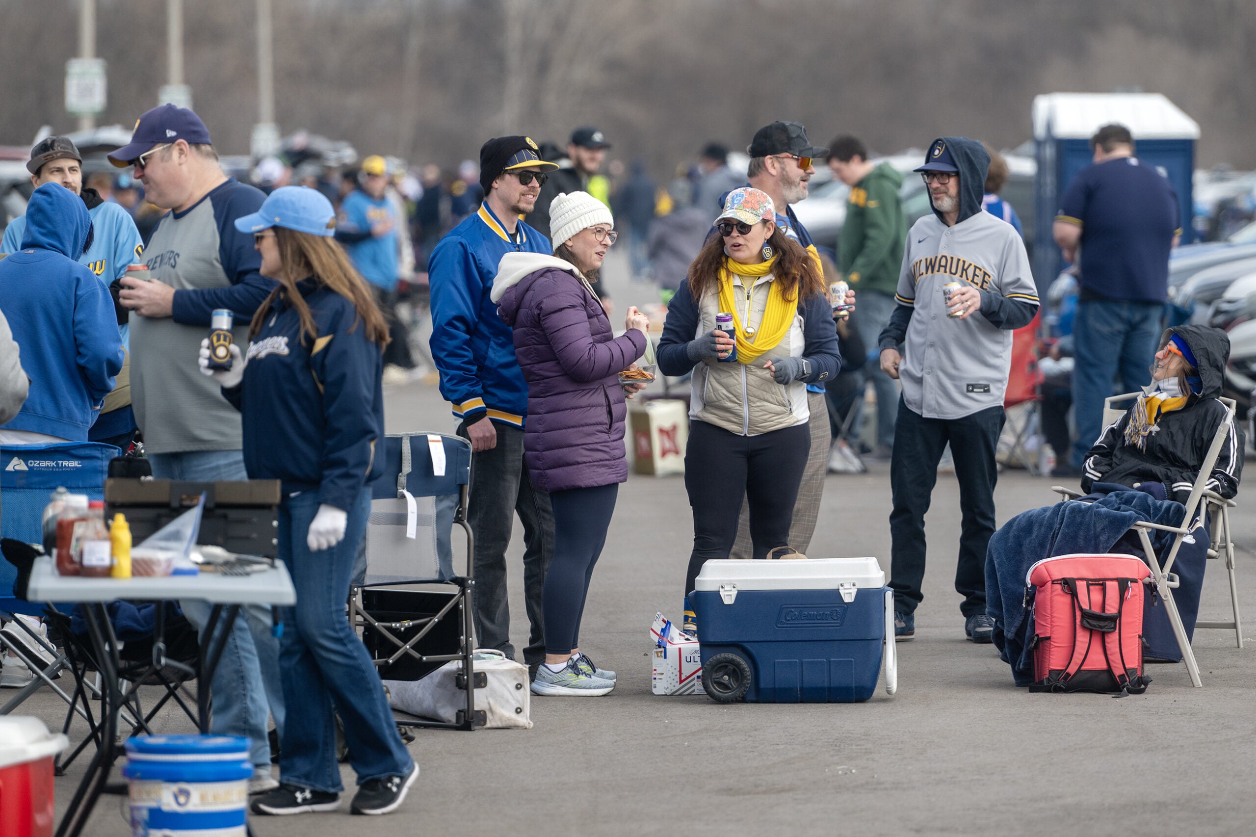A group of people in jackets and baseball gear stand and sit around coolers and a folding table at a parking lot tailgate.