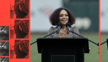 A woman with curly hair stands at a podium labeled "Oracle Park," speaking into two microphones, with red and black baseball-themed images on the left.