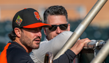 A man in a San Francisco Giants cap and jersey leans on a railing while talking to a man in sunglasses behind him.
