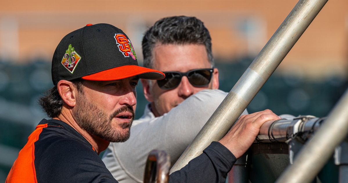 A man in a San Francisco Giants cap and jersey leans on a railing while talking to a man in sunglasses behind him.