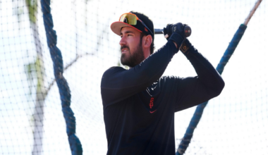 A man wearing a black long-sleeve shirt, black gloves, and a baseball cap holds a bat, preparing to swing inside a batting cage.