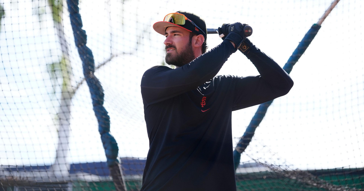 A man wearing a black long-sleeve shirt, black gloves, and a baseball cap holds a bat, preparing to swing inside a batting cage.