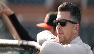 A man wearing sunglasses leans on a baseball dugout railing, looking intently at the field, with another person holding a bat blurred in the background.