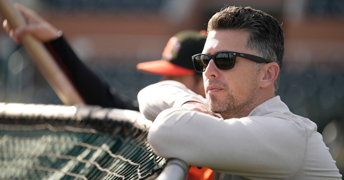 A man wearing sunglasses leans on a baseball dugout railing, looking intently at the field, with another person holding a bat blurred in the background.