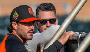 A man in a San Francisco Giants cap and jersey leans on a railing while talking to a man in sunglasses behind him.