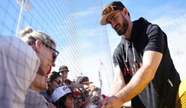 A baseball player wearing a Giants cap and sunglasses signs a ball for fans behind a fence on a sunny day.