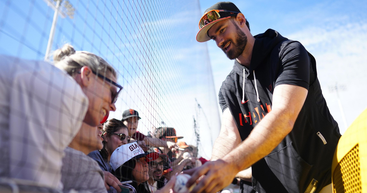 A baseball player wearing a Giants cap and sunglasses signs a ball for fans behind a fence on a sunny day.