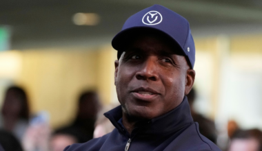 A man wearing a navy blue cap and zip-up jacket looks slightly to the side with a calm expression, while blurred people fill the background.