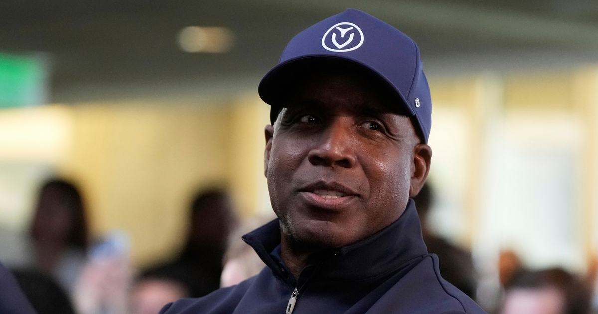 A man wearing a navy blue cap and zip-up jacket looks slightly to the side with a calm expression, while blurred people fill the background.