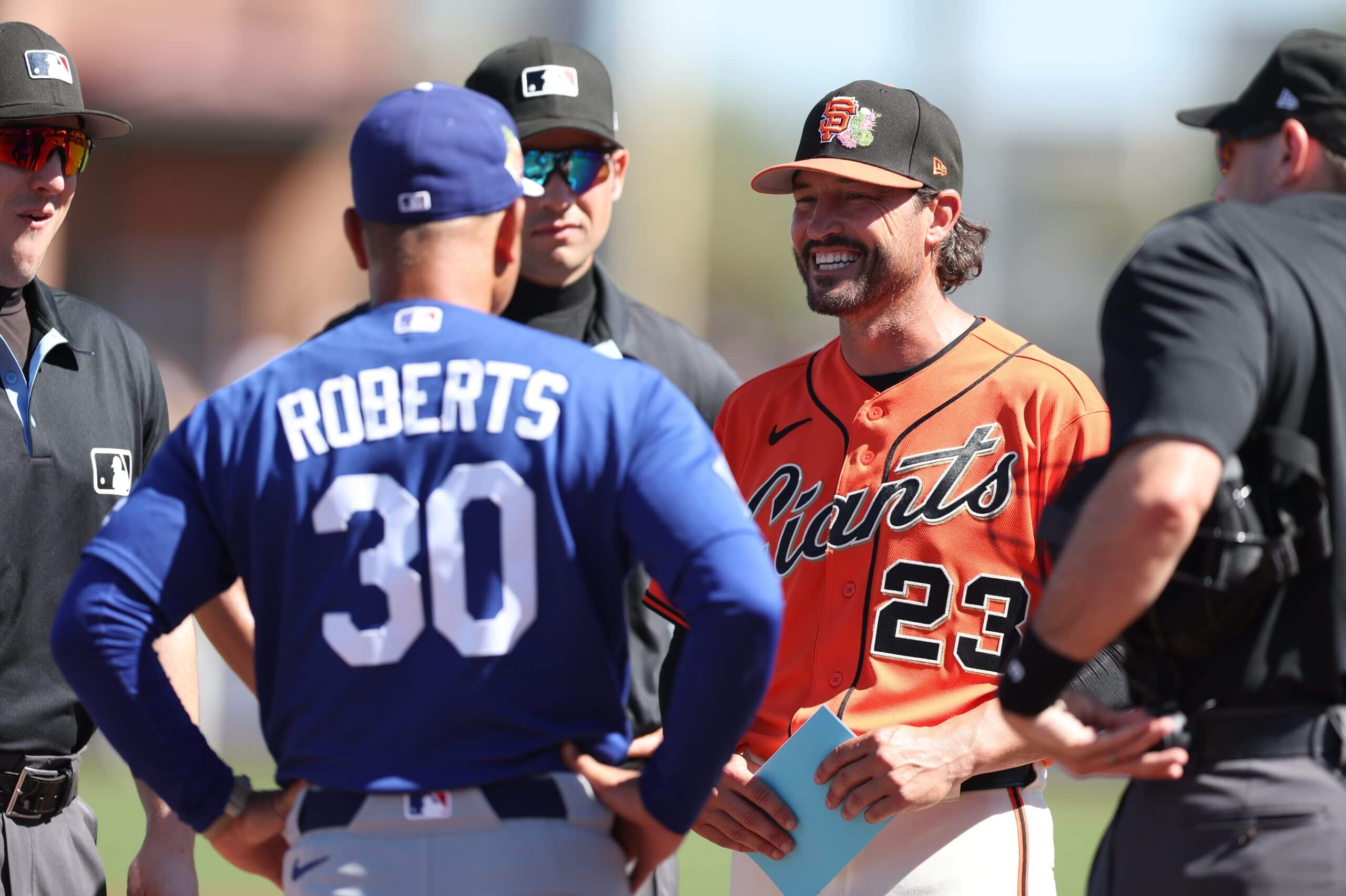 An image of San Francisco Giants manager Tony Vitello smiling as he huddles with MLB umpires and Los Angeles Dodgers manager Dave Roberts. 