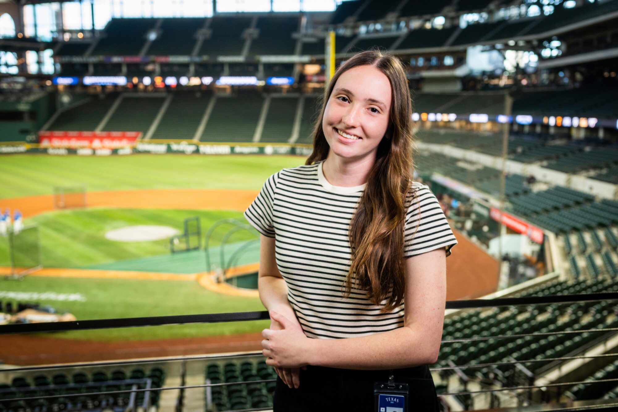 Ruby Blaylock smiles for the camera with the Texas Rangers stadium behind her. She is up in the seating area with the playing field behind her.