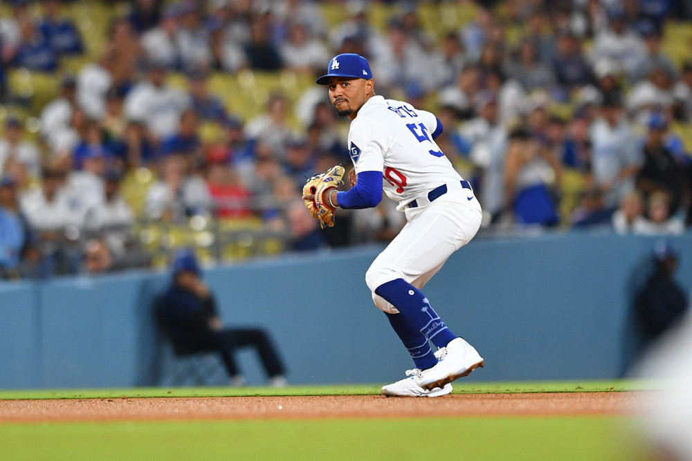 Mookie Betts, wearing a white Dodgers jersey with blue Betts written across the back and a blue cap with the Dodgers logo, prepares to make a throw to first base.