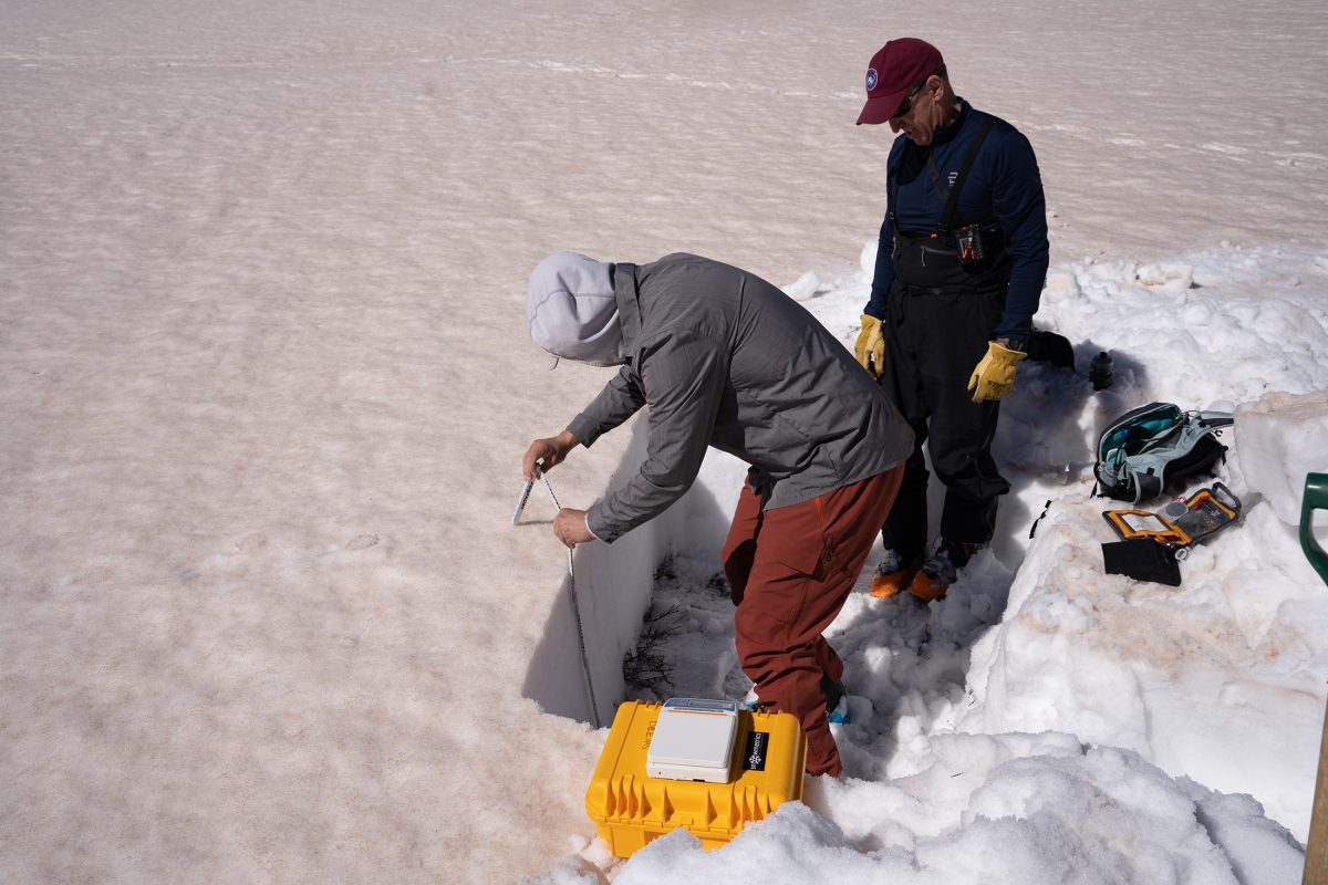 Two people in winter clothing carved out a section of snow, creating a cross section from the top of the snow to the ground. One holds up a tool to measure the height. Scientific tools are scattered around.