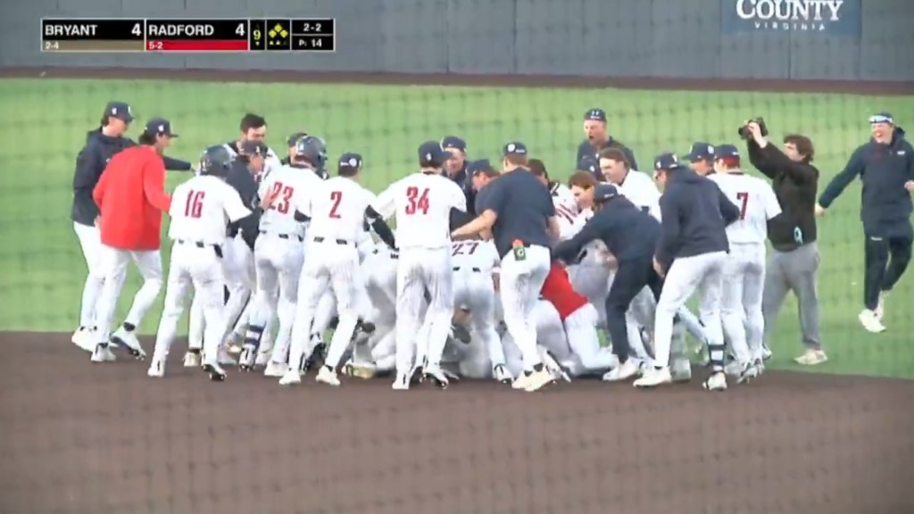 A large group of college baseball players in white uniforms and dark caps pile on top of each other at the end of a game.