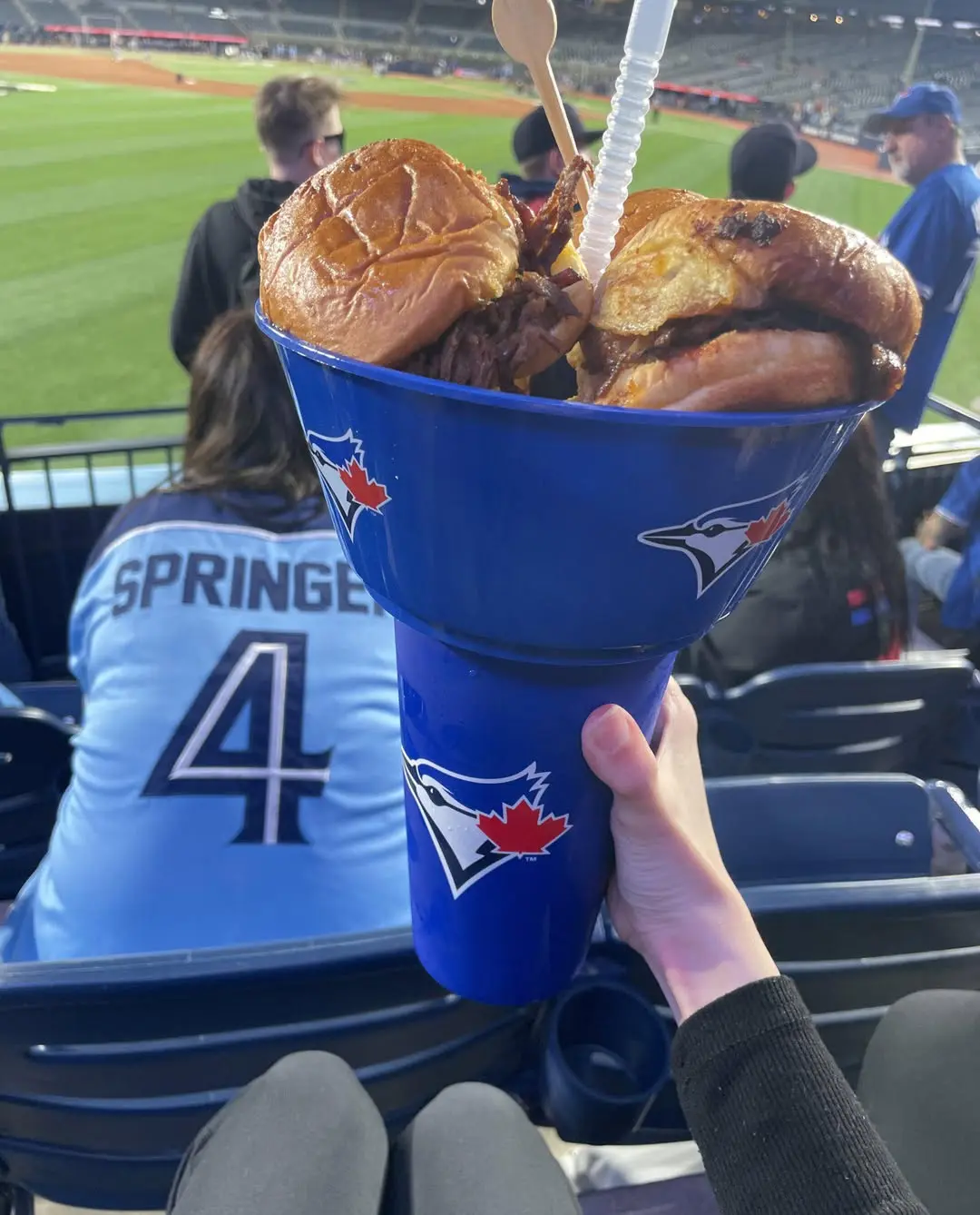 A hand holding a blue Jays-themed cone filled with burgers and fries during a game at Rogers Centre, with fans and the baseball field in the background.