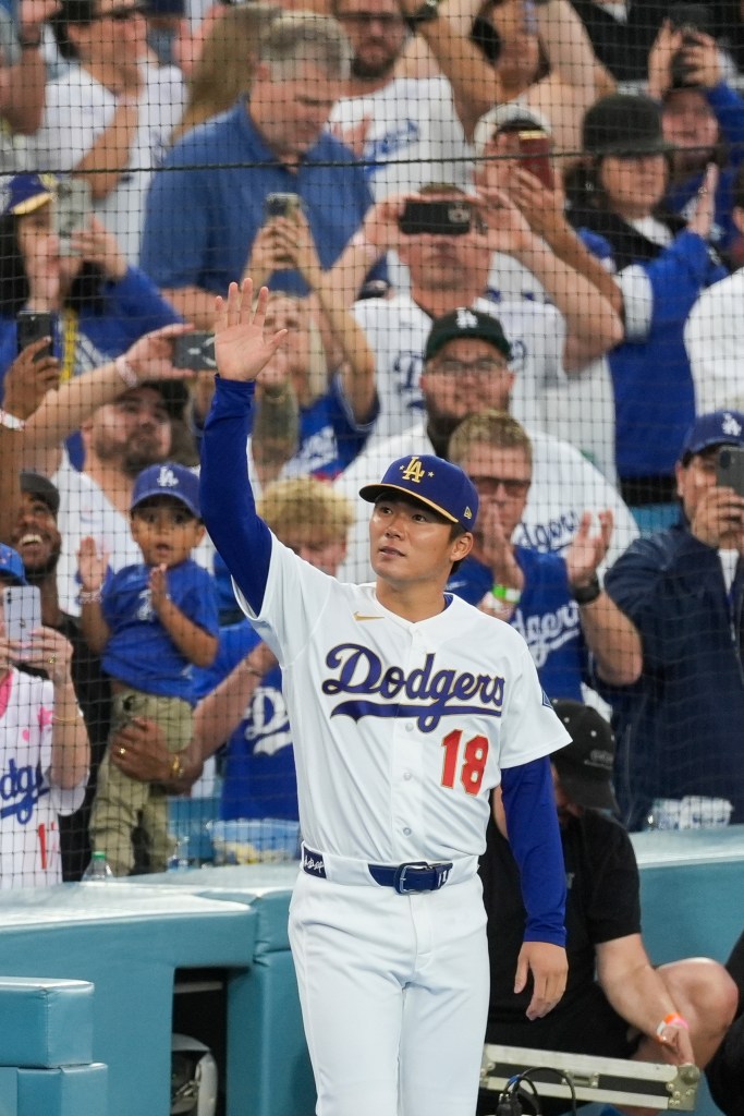 Los Angeles Dodgers player Yoshinobu Yamamoto waves to fans while wearing his jersey with the number 18 and a blue cap.