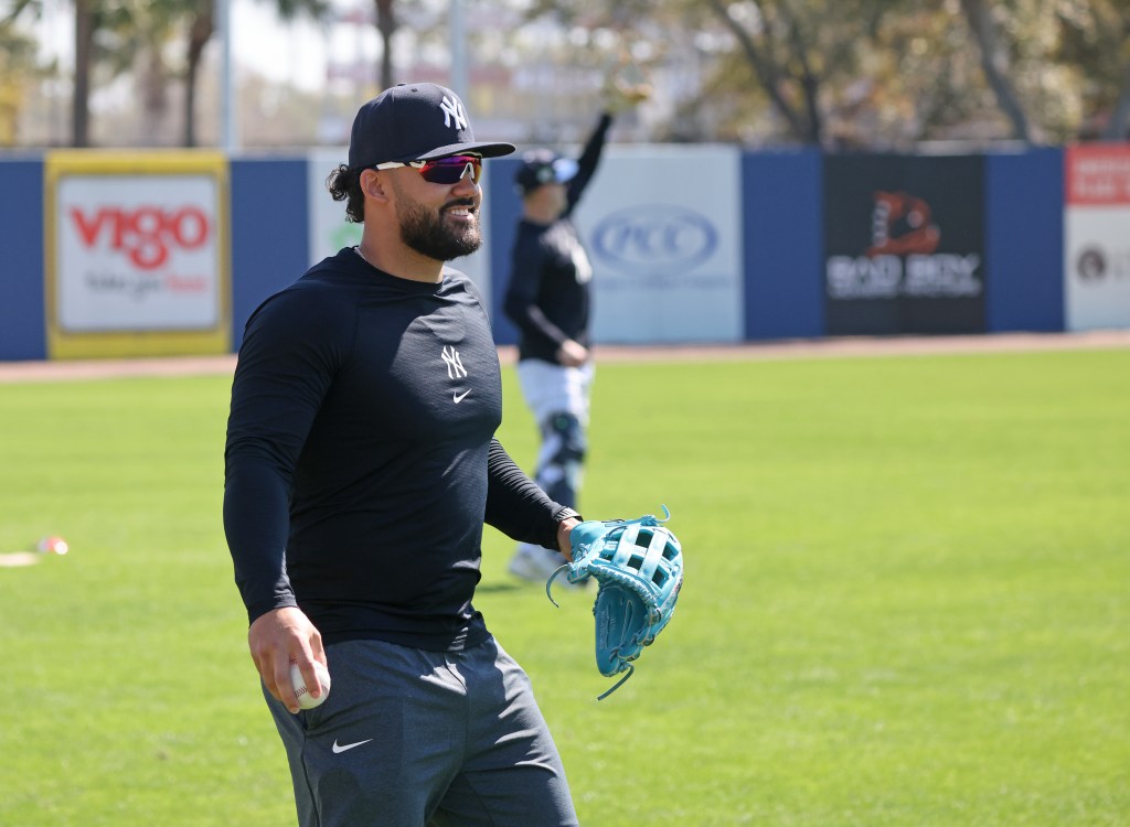 Jasson Domínguez smiling on the field in a Yankees cap and long-sleeve shirt, holding a baseball in one hand and a blue glove in the other.