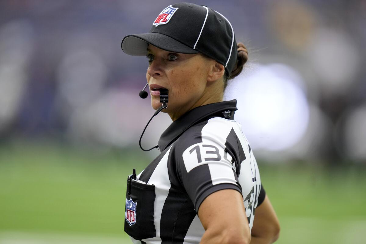 A referee holds a whistles in her mouth and looks to the left as she stands onfield. She wears a black cap with the NFL logo