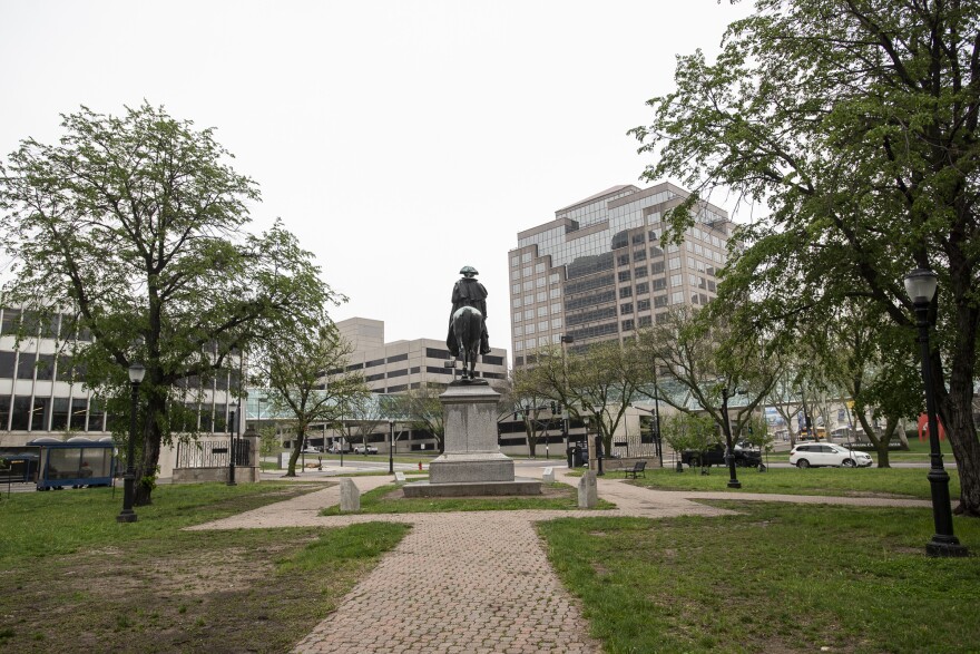 A large green space shows many trees  on either side of the frame. Many large office buildings can be seen in background. The rear of a bronze statue of George Washington riding a horse is in the middle of the frame.