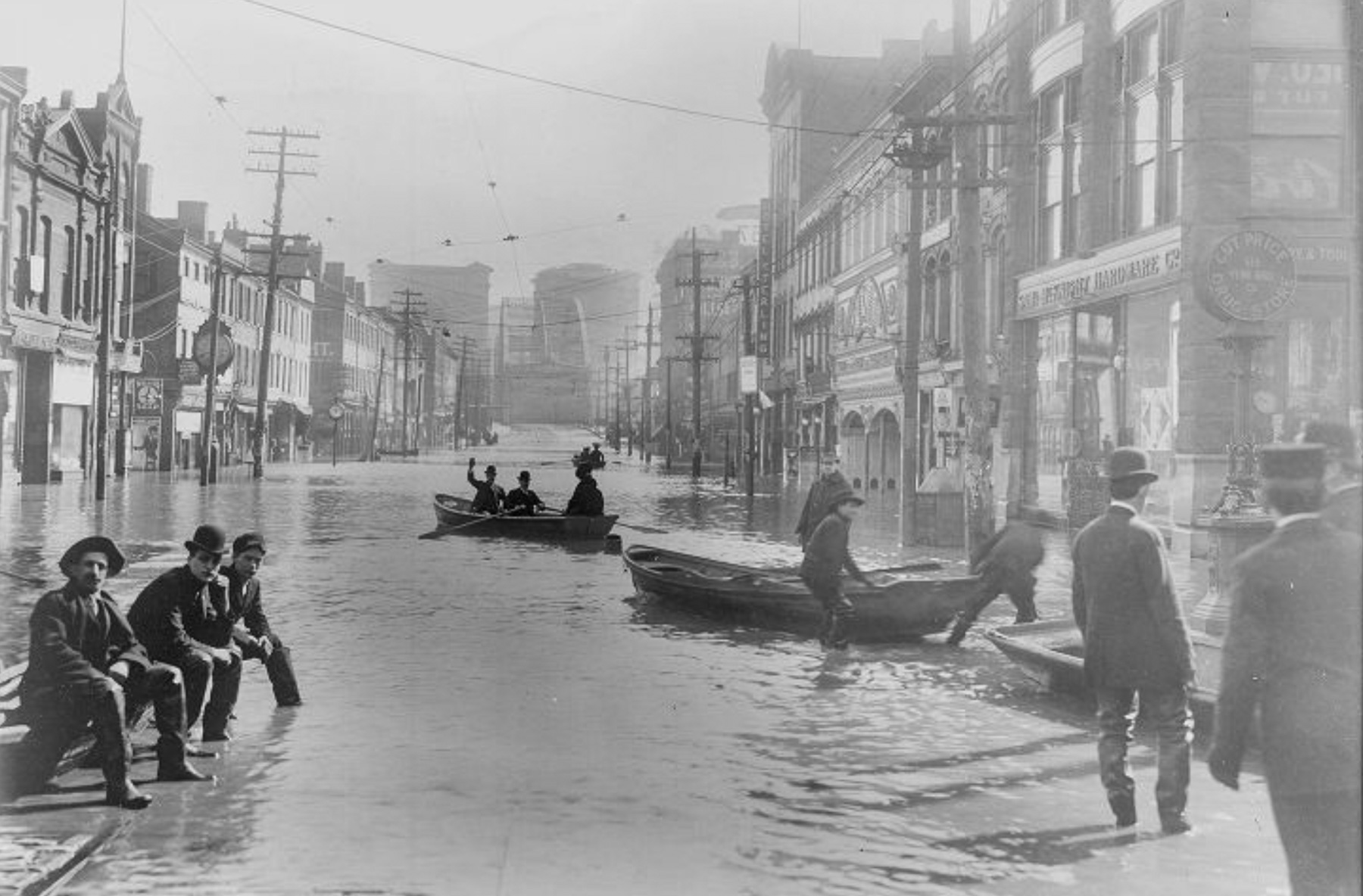 Black and white photo of a flooded city street with people sitting on ledges and others navigating the water in rowboats. Buildings and utility poles line the street.