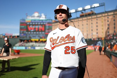 Pete Alonso in white Orioles uniform wearing headset in front of dugout with Os Warehouse behind him