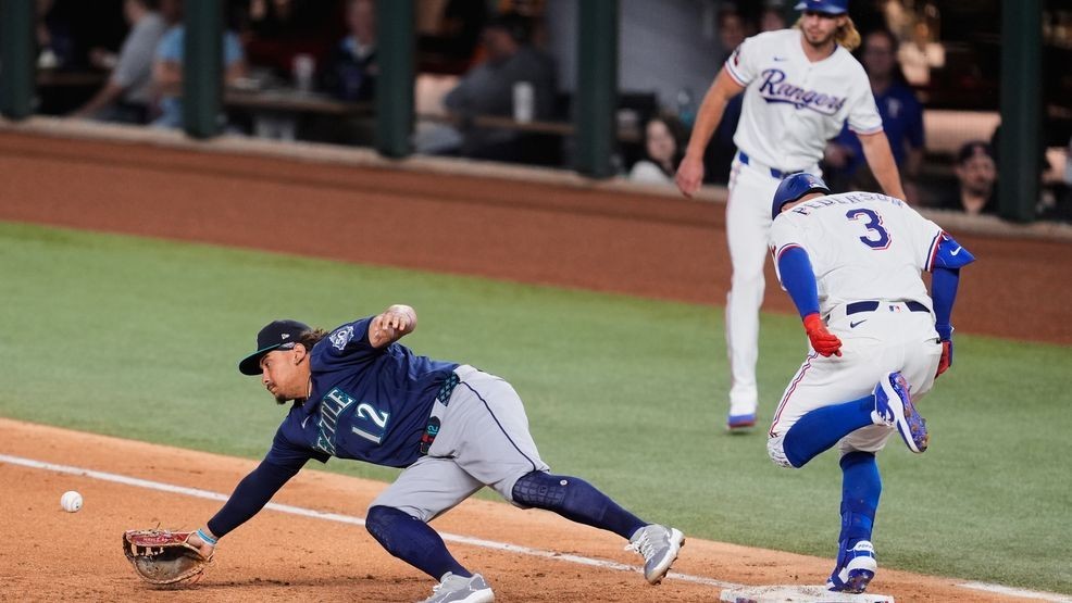 Texas Rangers' Joc Pederson (3) reaches on a single as Seattle Mariners first baseman Josh Naylor (12) dives for an errant throw to the bag in the fifth inning of a baseball game Tuesday, April 7, 2026, in Arlington, Texas. (AP Photo/Tony Gutierrez)