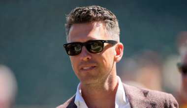 A man with short, styled hair wears black sunglasses, a white collared shirt, and a maroon blazer, standing outdoors in soft sunlight.