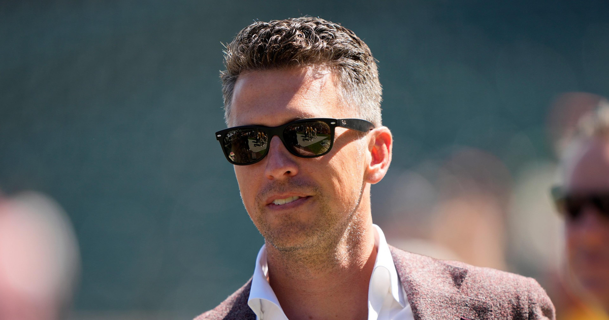 A man with short, styled hair wears black sunglasses, a white collared shirt, and a maroon blazer, standing outdoors in soft sunlight.