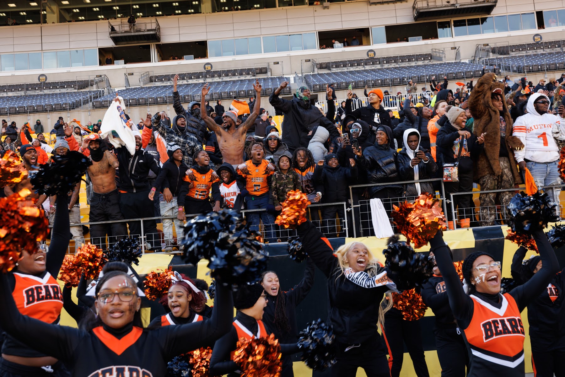 A group of cheerleaders performs in front of an enthusiastic crowd of fans, some shirtless and cheering, in a stadium setting.
