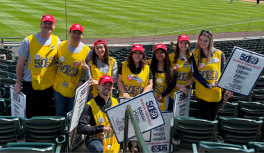 Group of volunteers in yellow vests and red hats pose in stadium seating while selling 50/50 raffle tickets at a Colorado Rockies game.