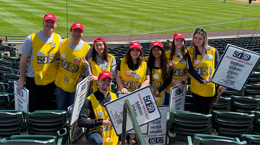 Group of volunteers in yellow vests and red hats pose in stadium seating while selling 50/50 raffle tickets at a Colorado Rockies game.