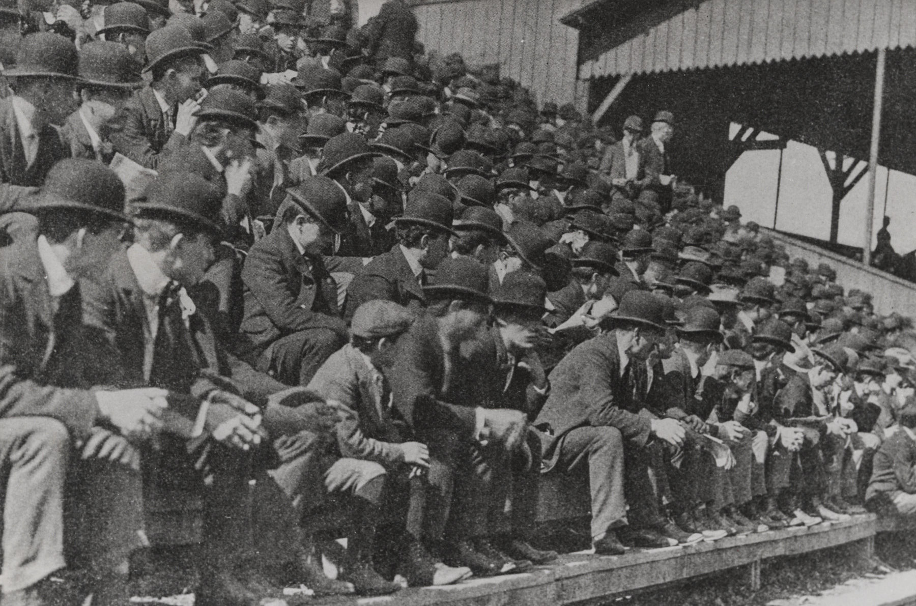 A large crowd of men in suits and bowler hats sit closely together on wooden bleachers at an outdoor event, possibly early 20th century.