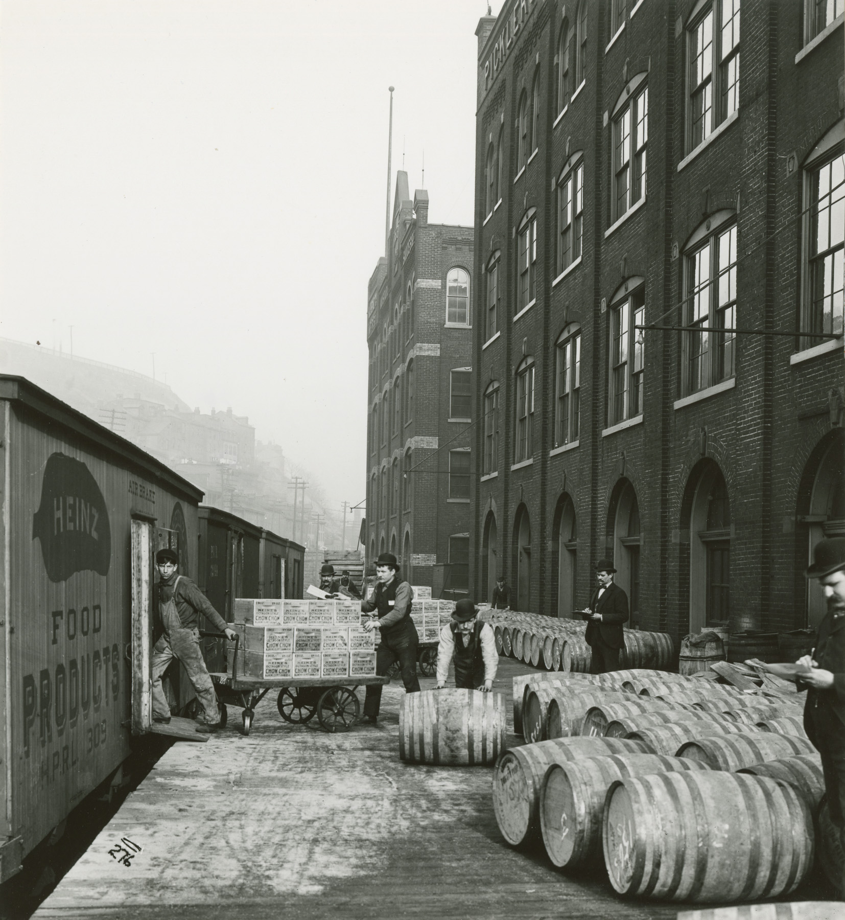 Workers unload boxes and barrels from a Heinz Food Products train car outside a brick factory building, stacking goods on the loading dock.