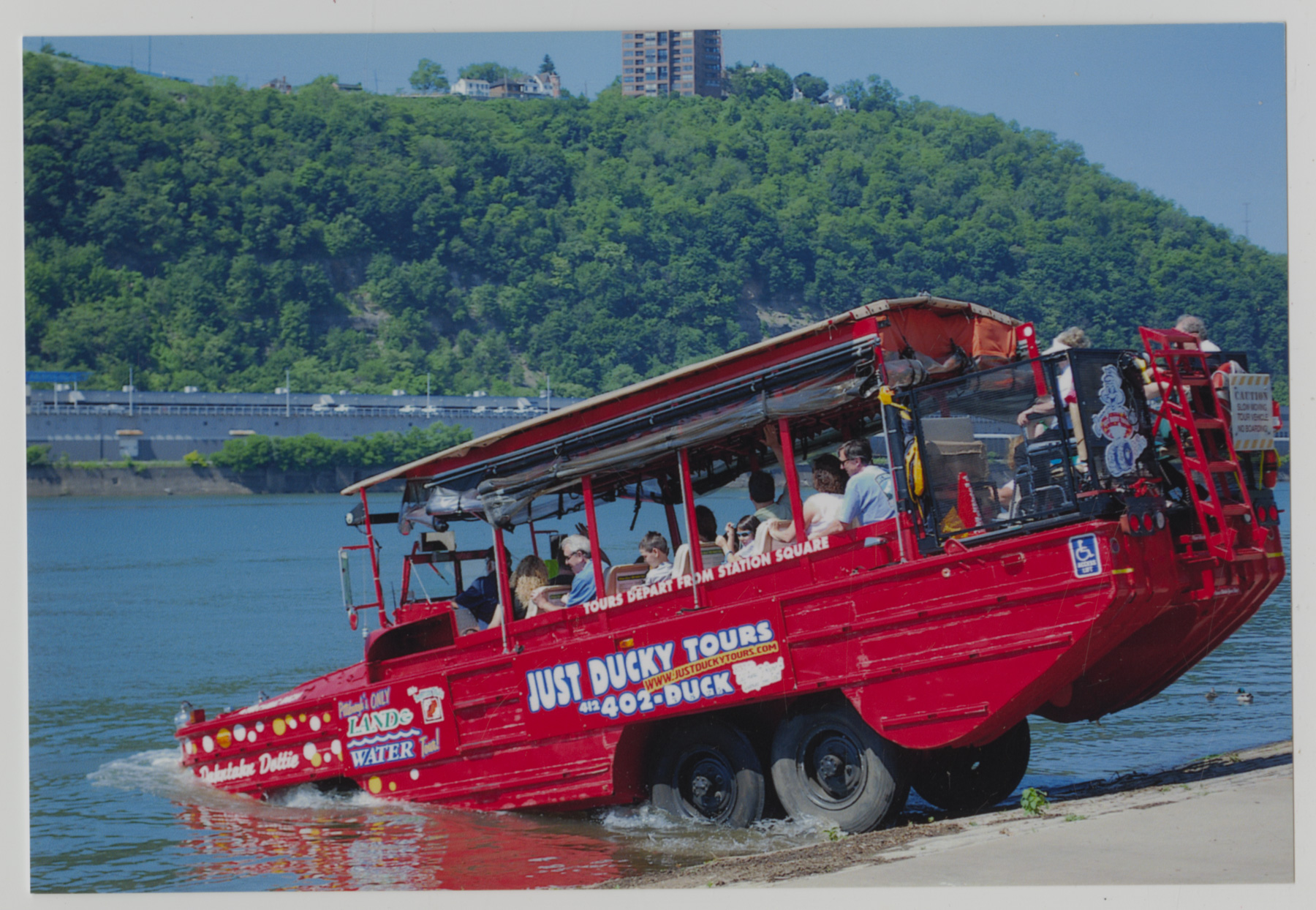 A red amphibious tour vehicle labeled "Just Ducky Tours" enters the water from a boat ramp, carrying passengers near a wooded hillside.