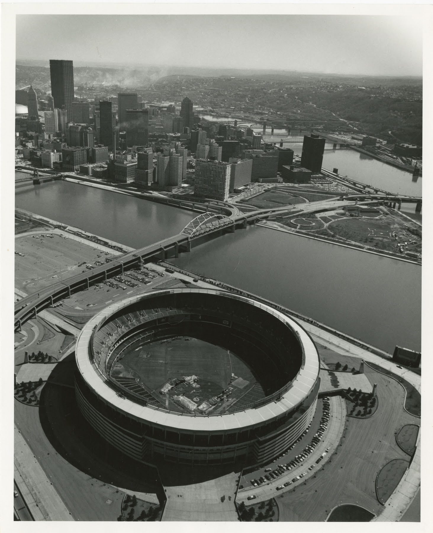 Aerial view of a large circular stadium next to a river with bridges, with a city skyline and riverbanks visible in the background.