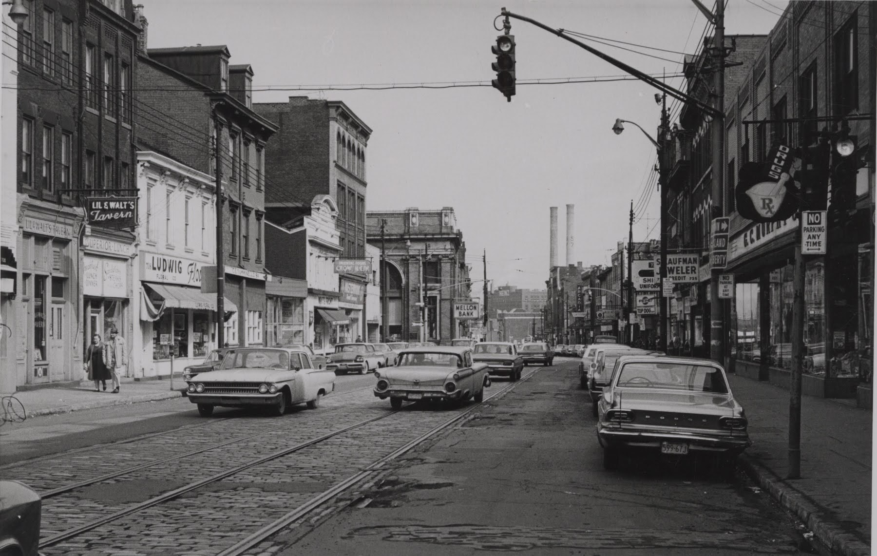 Black-and-white photo of a city street with parked cars, old buildings, shops, a traffic light, and industrial smokestacks in the distance.