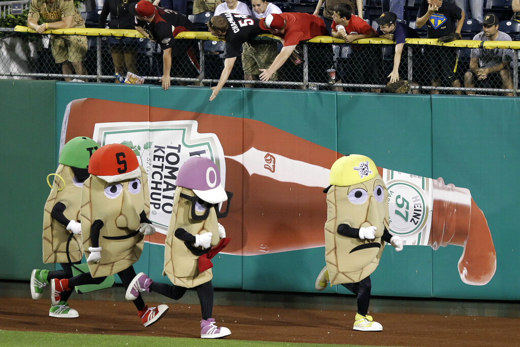 Four people in condiment-themed mascot costumes run a race on a baseball field, while fans reach over the outfield wall above a large ketchup bottle advertisement.