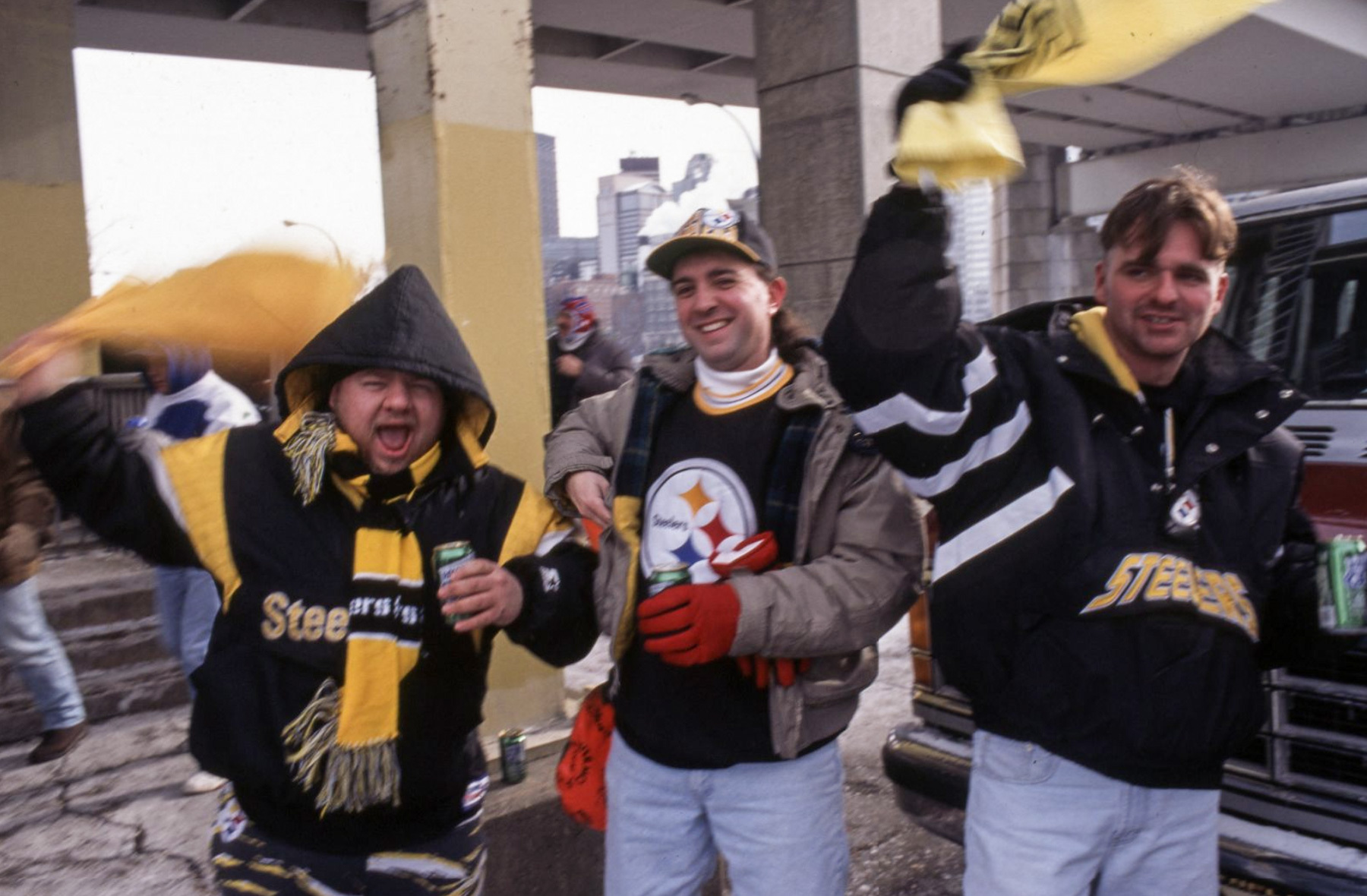 Three people dressed in Pittsburgh Steelers gear cheer and wave yellow towels outdoors near a vehicle, with city buildings visible in the background.