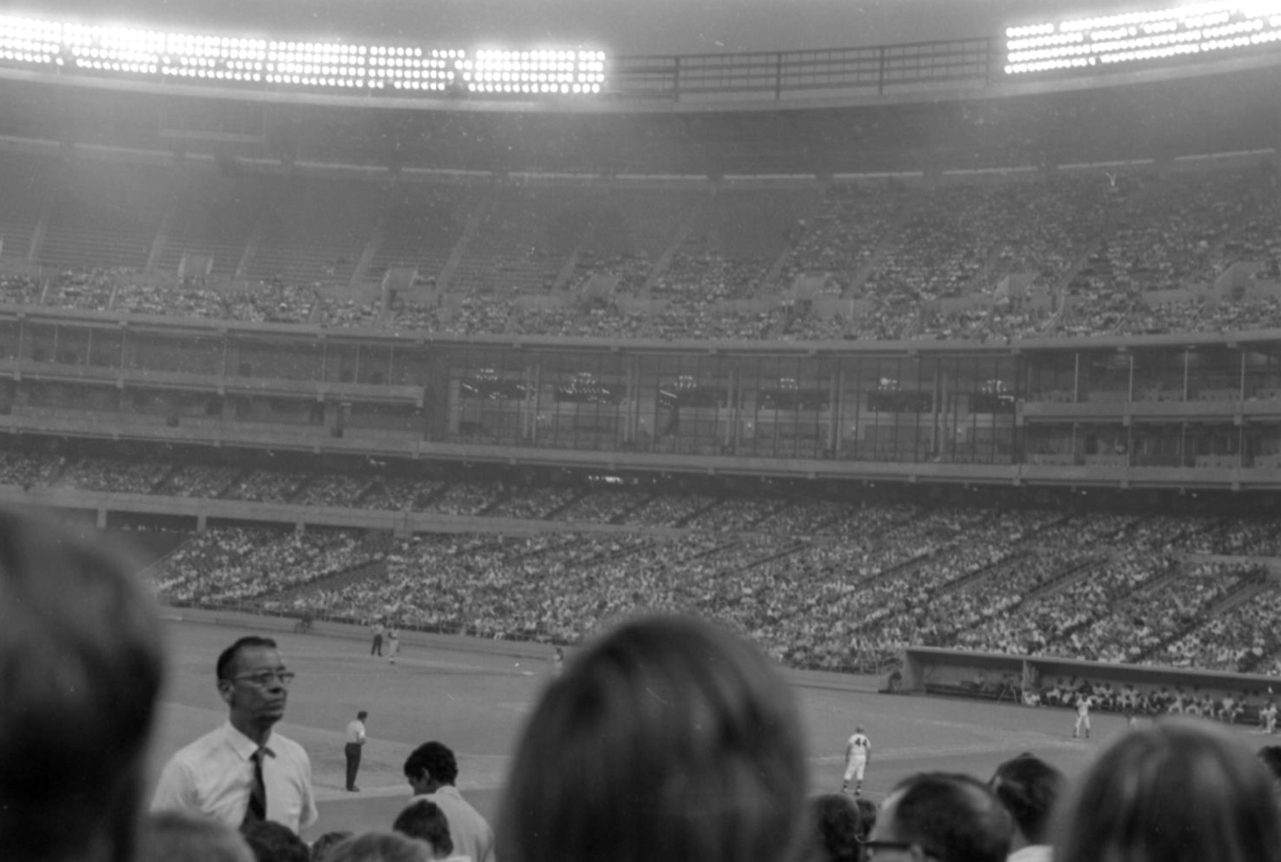 A black-and-white photo of a baseball stadium filled with spectators. Several players are on the field, and a man in glasses stands in the foreground among the crowd.