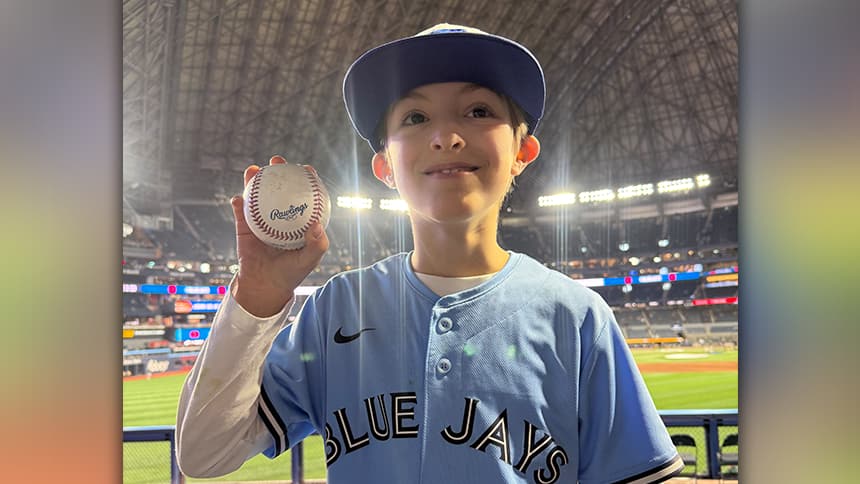 A boy wearing a Blue Jays jersey holding a baseball in his hand as he smiles to the camera.