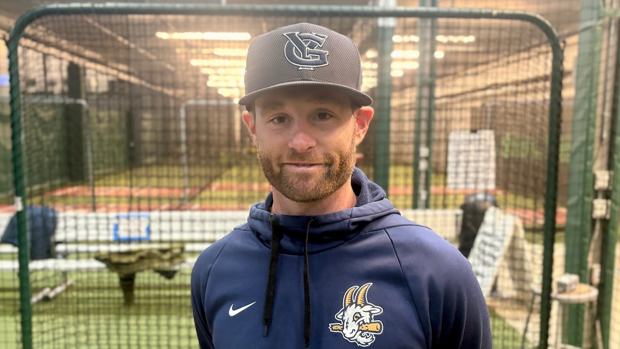 A man in a ball cap poses for a photo in front of indoor batting cages.