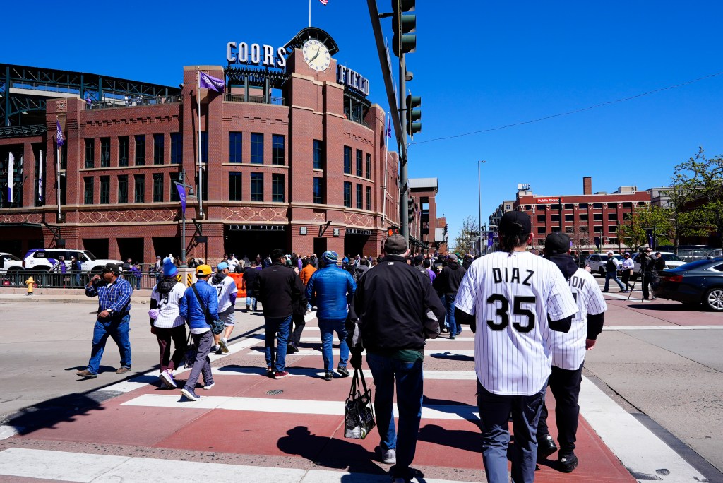People walking towards Coors Field, a large brick baseball stadium, on a sunny day.