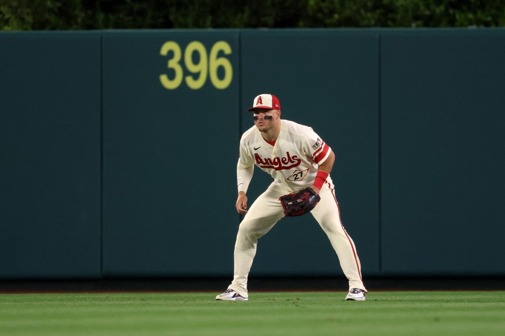 Baseball player Mike Trout standing on the field in a ready position, wearing a cream and red Angels uniform with number 27, in front of a dark green outfield wall with the number 396.