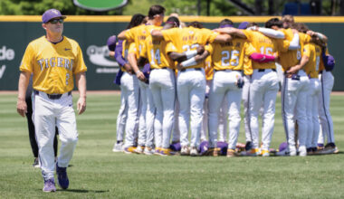 Baseball team in yellow jerseys huddled together on the field; a coach walks in the foreground.