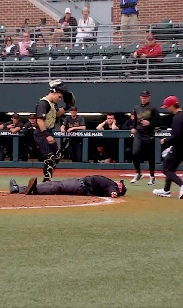 A baseball umpire lies face down on the ground behind home plate, with a catcher standing nearby and other players in the dugout.